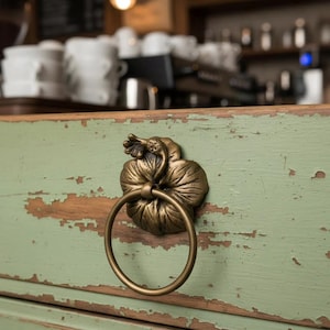 May include: A close-up of a bronze-colored drawer pull in the shape of a flower with a ring handle. The drawer is painted a distressed green color, with visible wear and tear. The background is blurred, suggesting a cafe setting.
