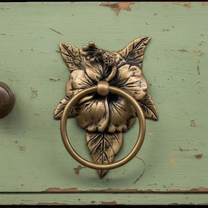 May include: Close-up of a bronze-colored floral drawer pull with a ring handle. The pull is shaped like a flower with detailed leaves and petals. It is mounted on a distressed, light green painted wooden surface.