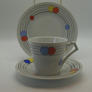 May include: A white ceramic tea cup and saucer set accompanied by a matching side plate. Each piece features a pattern of thin black concentric lines accented with small, solid circles in primary colours of red, yellow, and light blue.
