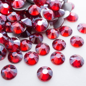 May include: A collection of faceted, round, red rhinestones with silver backing. The gemstones are scattered across a white surface, catching the light and displaying a brilliant sparkle. These embellishments are ideal for various craft projects.