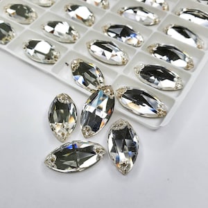Puede incluir: Primer plano de un grupo de piedras de cristal transparentes, facetadas, en forma de marquesa. Las piedras de cristal están dispuestas en un grupo suelto sobre una superficie blanca.