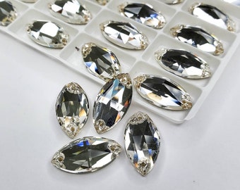 May include: A close-up of a group of clear, faceted, marquise-shaped rhinestones. The rhinestones are arranged in a loose cluster on a white surface.