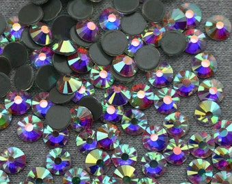 May include: A large quantity of iridescent AB crystal rhinestones, also known as glass gems, in various sizes. The rhinestones are scattered on a gray fabric background.