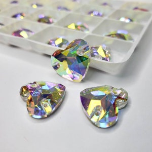 May include: Iridescent heart-shaped crystal beads with a faceted design. The beads are in a white tray with individual compartments. The crystals reflect a rainbow of colors, including yellow, green, and purple. The beads have a hole for stringing.