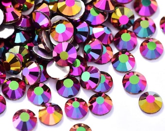 Puede incluir: Un montón de piedras de cristal de color rosa, verde y dorado iridiscente. Las piedras de cristal están esparcidas sobre una superficie blanca.