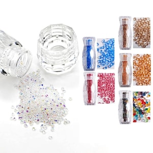 Puede incluir: Contenedores de vidrio transparente y facetado y pedrería iridiscente dispersa. Varios frascos pequeños contienen pedrería colorida en azul, rosa, dorado y multicolor. Ideal para arte de uñas o proyectos de manualidades.