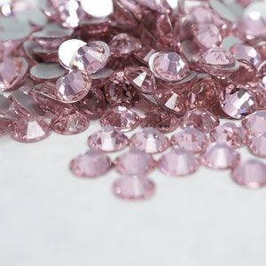 Puede incluir: Un montón de piedras de cristal de color rosa, también conocidas como cristales o gemas, esparcidas sobre una superficie blanca. Son pequeñas, facetadas y tienen un aspecto brillante y brillante.