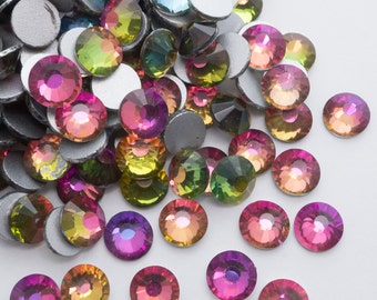 May include: Assortment of iridescent glass rhinestones in various colors, including pink, green, blue, and yellow. The rhinestones are scattered on a white surface.