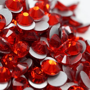 May include: A close-up of a pile of red rhinestones with silver backing. The rhinestones are faceted and have a shiny finish.