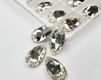 May include: Clear, faceted teardrop-shaped rhinestones. Some rhinestones are arranged in a white tray, while others are scattered on a white surface. The rhinestones have a silver backing and are designed with multiple facets to reflect light.
