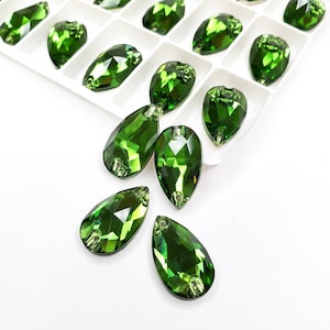 Puede incluir: Un grupo de piedras de cristal verde perlado en forma de lágrima, algunas están en un contenedor de plástico blanco, otras están esparcidas sobre una superficie blanca.