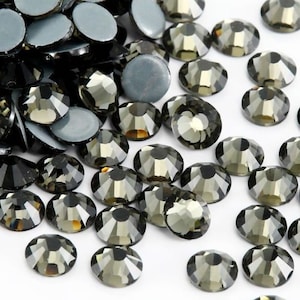 May include: A large quantity of dark gray rhinestones, also known as crystals or gems, are scattered on a white surface. The rhinestones are round and have a faceted design.