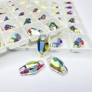 May include: Assortment of iridescent glass rhinestones in a white plastic container. The rhinestones are oval shaped with two holes at the top.