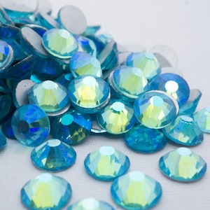 Op de afbeelding: Een stapel lichtblauwe, iriserende strasssteentjes, ook wel glazen edelstenen genoemd, met een regenboogkleureffect. De strasssteentjes zijn verspreid over een wit oppervlak.