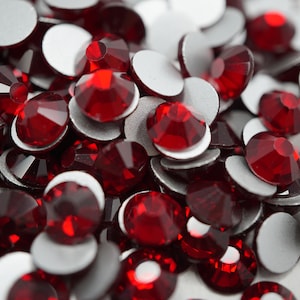 May include: A close-up of a pile of red rhinestones with adhesive backing. The rhinestones are round and have a faceted design.