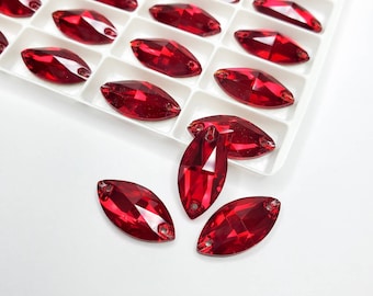 May include: Marquise-shaped, faceted red glass rhinestones with two holes for sewing or gluing. The rhinestones are displayed in a white plastic tray and scattered on a white surface. These decorative embellishments are ideal for crafting and jewelry making.
