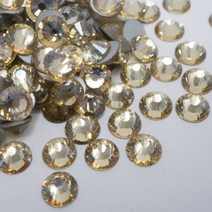 May include: A large quantity of small, round, clear glass rhinestones with a light gold shimmer. The rhinestones are scattered on a white surface.