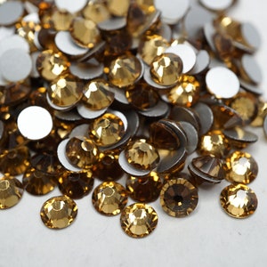 May include: A pile of gold-coloured rhinestones with adhesive backs. The rhinestones are scattered on a white surface.
