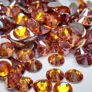 May include: A large quantity of orange iridescent rhinestones, also known as glass crystals, are scattered on a white surface. The rhinestones have a faceted design and a shiny, reflective finish.