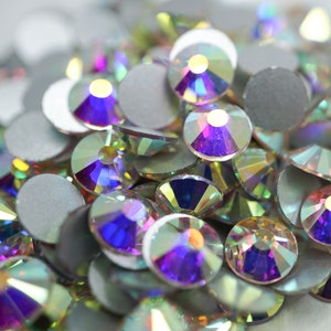 May include: A close-up of iridescent glass rhinestones in various shades of purple, blue, and green. The rhinestones are scattered on a white surface.