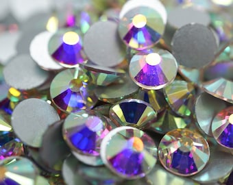 May include: A close-up of iridescent glass rhinestones in various shades of purple, blue, and green. The rhinestones are scattered on a white surface.