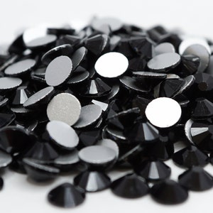 May include: A pile of black rhinestones with silver backs. The rhinestones are scattered on a white surface.