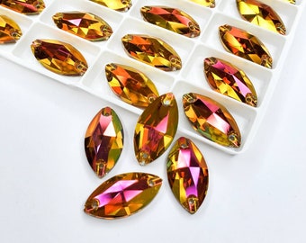 May include: Marquise-shaped iridescent rhinestones in a white tray. The rhinestones have a faceted surface that reflects a spectrum of colors, including gold, orange, and pink. Each stone has two holes for sewing or attaching. The tray is divided into individual compartments.