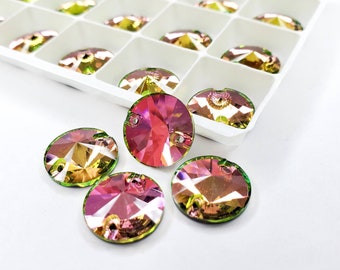 May include: Iridescent, faceted crystal rhinestones in shades of pink, green, and gold. Several round rhinestones are scattered in front of a white tray filled with similar stones. Each stone has two small holes.