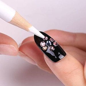 Puede incluir: Un bolígrafo blanco para decoración de uñas se utiliza para aplicar pegamento a una uña negra con un grupo de piedras de cristal de colores.