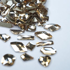 Puede incluir: Una colección de pedrería de color ámbar de varias formas, dispersas sobre una superficie blanca. Las formas incluyen corazones, estrellas, rectángulos y formas geométricas. Algunas piedras tienen un aspecto facetado.