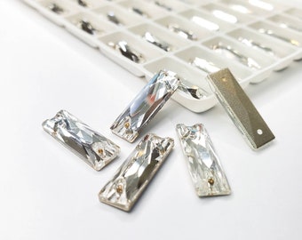 May include: Close-up of several clear, rectangular crystal rhinestones with facets, scattered on a white surface. Some rhinestones are in a white plastic organizer. The crystals have small holes for sewing or gluing, ideal for embellishments.