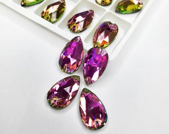 May include: A group of iridescent purple teardrop-shaped rhinestones with a green and gold shimmer. The rhinestones are scattered on a white surface.