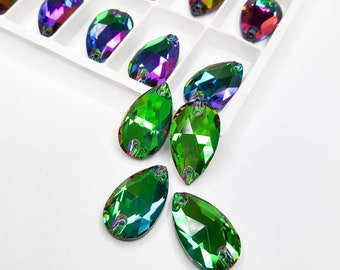 May include: A collection of iridescent teardrop-shaped crystals in shades of green, purple, and blue. Some crystals are arranged in a white tray, while others are scattered on a white surface. These faceted crystals have a hole at the top.