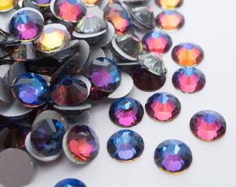 May include: A collection of iridescent, round rhinestones with a flat back. The gems display a spectrum of colors, including blue, purple, red, and yellow, with a metallic silver backing. These decorative embellishments are scattered on a white surface.