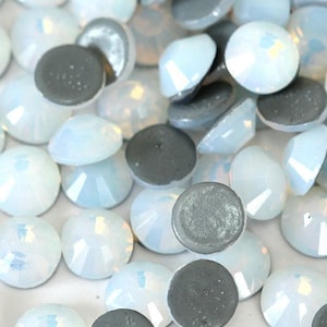 Puede incluir: Primer plano de un montón de piedras de cristal de opal blanco. Las piedras de cristal son redondas y tienen una superficie facetada. Están esparcidas sobre una superficie blanca.