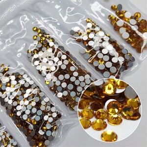 Puede incluir: Varias bolsas de pequeños diamantes de imitación redondos de color ámbar con respaldo plateado. Los diamantes de imitación están dispuestos en filas en las bolsas. Una imagen de primer plano muestra los diamantes de imitación esparcidos sobre una superficie blanca.