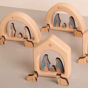 Op de afbeelding: Een verzameling van vier houten kerststalfiguren. Elke scène toont een centrale boog of huisvorm met figuren van Maria, Jozef en het kindje Jezus. De figuren zijn gemaakt van lichtgekleurd hout met geschilderde details en kleine dierenfiguren.