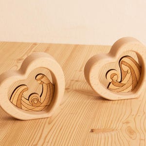 Può includere: Due ornamenti a forma di cuore in legno. Ogni ornamento presenta un design ritagliato che raffigura una scena religiosa. Le scene sono scolpite in una tonalità di legno a contrasto e inserite all'interno della cornice del cuore. Gli ornamenti sono esposti su una superficie di legno chiaro.