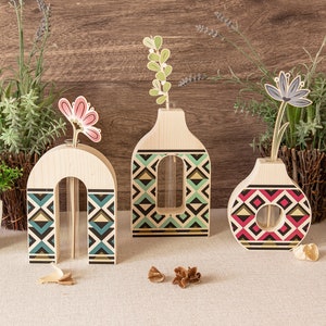 Könnte beinhalten: Drei Holzvasen mit geometrischen Mustern in Blau, Grün und Rot. Jede Vase hat ein klares Glasrohr zum Halten von Blumen. Die Vasen sind auf einer hellbraunen Oberfläche angeordnet.