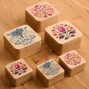Op de afbeelding: Zes kleine, vierkante houten dozen met bloemmotieven op de deksels. De dozen zijn gemaakt van lichtgekleurd hout en hebben kleurrijke, ingewikkelde bloempatronen in rood, roze, blauw en groen. De dozen zijn gerangschikt op een houten oppervlak.