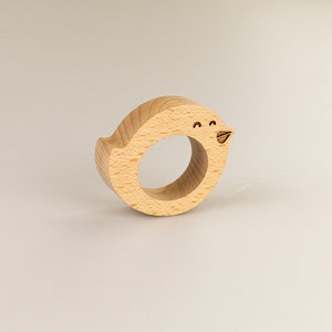 May include: A wooden bird-shaped napkin ring with a simple, hand-drawn face. The ring is made of light brown wood and is a fun and whimsical way to add a touch of personality to your table setting.