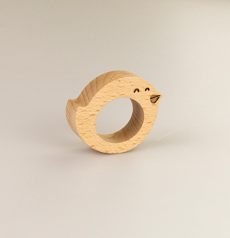 May include: A wooden bird-shaped napkin ring with a simple, hand-drawn face. The ring is made of light brown wood and is a fun and whimsical way to add a touch of personality to your table setting.