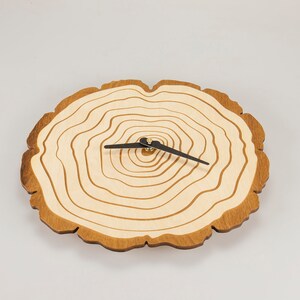 Pu&ograve; includere: Un orologio in legno con un design ad anelli di albero. Il quadrante dell'orologio &egrave; in legno chiaro con bordi marrone scuro. Le lancette dell'orologio sono nere.