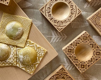 May include: Four wooden square molds with intricate designs and a round indentation in the center. The molds are used for making ravioli pasta. The molds are on a light brown surface with a beige fabric background.