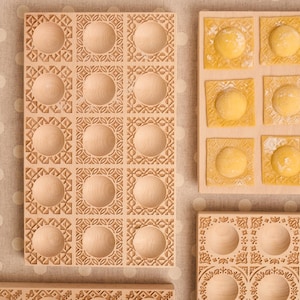 Può includere: Stampi per ravioli in legno con motivi decorativi. Uno stampo è riempito di ravioli freschi. Gli stampi hanno una forma quadrata con un'indentazione circolare al centro di ogni quadrato. Gli stampi sono in legno chiaro.