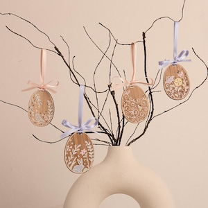 Peut inclure: Quatre œufs de Pâques en bois avec des motifs floraux et de lapin sont suspendus à une branche dans un vase. Les œufs sont peints dans des couleurs pastel et présentent des détails délicats.