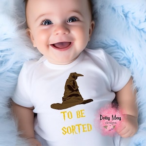 May include: A white short-sleeved baby bodysuit featuring a brown wizard hat graphic centered on the chest. Below the hat, the text reads "TO BE SORTED" in a yellow, jagged font. The garment has a round neckline and simple hemmed sleeves.