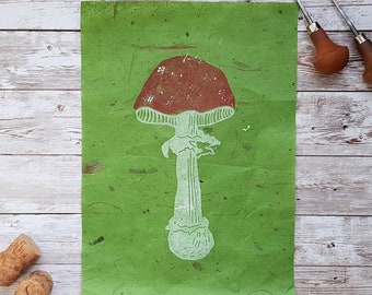 May include: A white mushroom with a red cap printed on green paper. The mushroom is a linocut print.