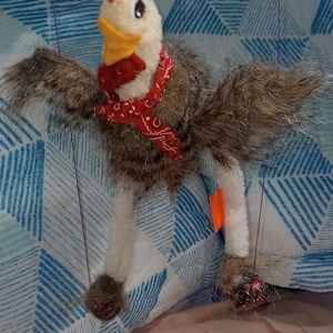 May include: A handmade chicken puppet with a white head, orange comb, and yellow beak. It has brown and gray feathered wings and a red bandana. The puppet is suspended by purple strings.