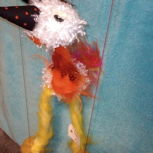 May include: A colorful felt puppet of a bird with a black and white polka dot beak, orange body, and yellow legs. The puppet is hanging from strings and has a price tag attached to one of its legs.